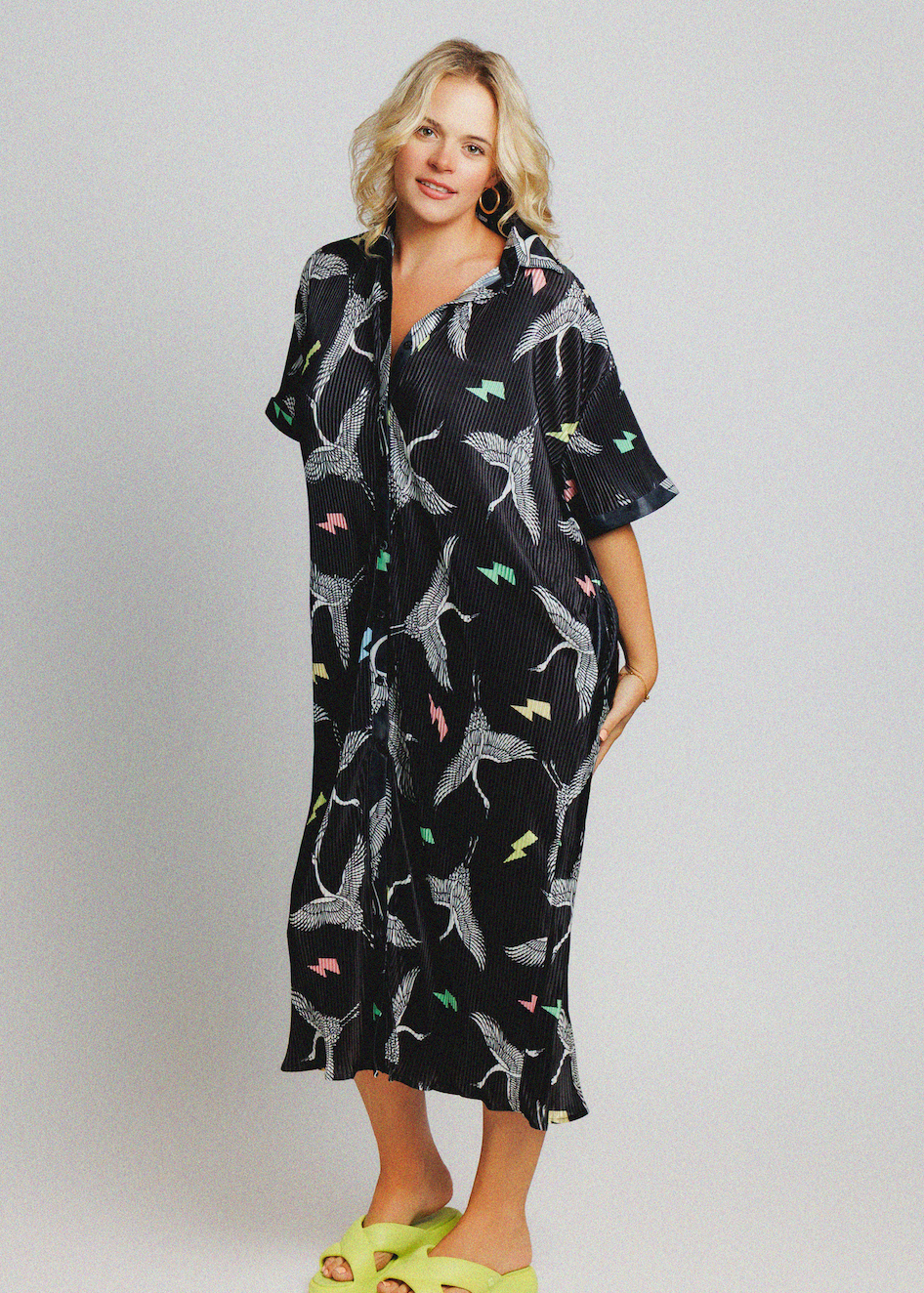 A woman modeling a black short-sleeve kaftan-style dress with a white shark print and small multicolored accents, paired with neon yellow slide sandals, against a light gray studio background.