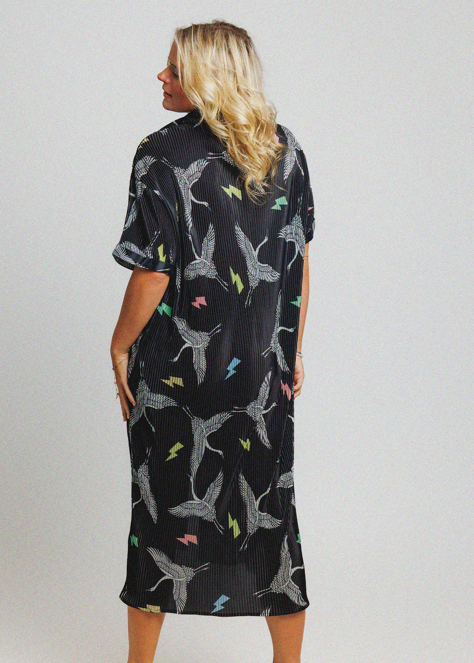 A woman viewed from the back wearing a short-sleeve black midi dress with a white bird print and small colorful accents, against a light gray studio background.