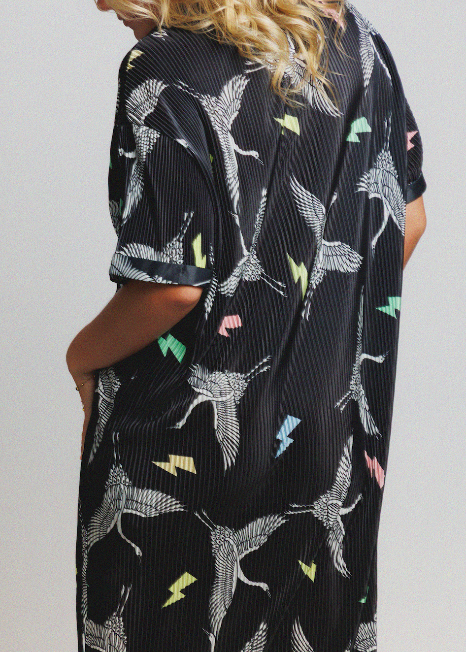 A woman wearing a black short-sleeve kaftan dress with a white bird print and colorful lightning bolt accents, shown from a side-back angle against a light gray background.