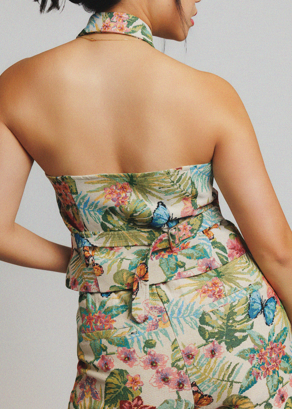 A close-up back view of a model wearing a strapless halter dress with a tropical floral and butterfly print on a light beige background.