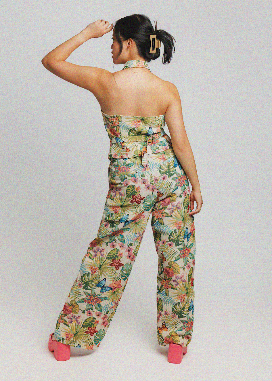 A woman is shown from behind wearing a strapless tropical floral print jumpsuit with wide-leg pants and a matching belt, paired with pink high heels, against a light gray studio background.