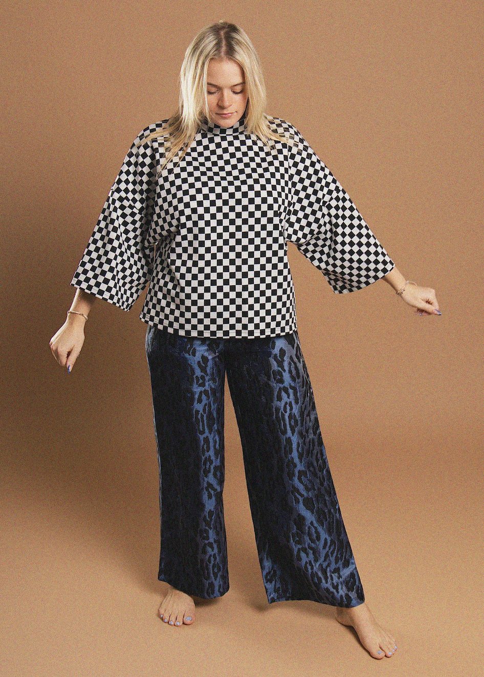 A woman modeling a black and white checkered top with wide three-quarter sleeves and dark blue leopard-print wide-leg pants, standing against a solid tan studio background.