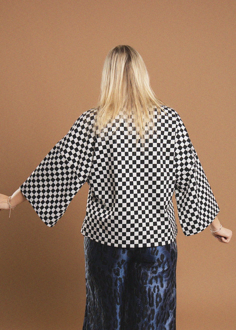 A woman viewed from behind wearing a black and white checkered top with wide bell sleeves and dark blue patterned pants, standing against a solid tan background.
