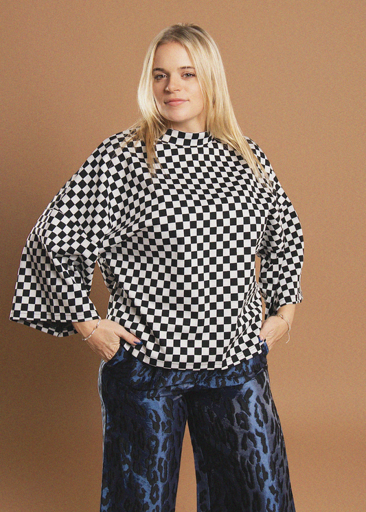 A woman modeling a black and white checkered blouse with wide flared sleeves and a high mock neck, paired with blue leopard-print pants, against a solid tan studio background.