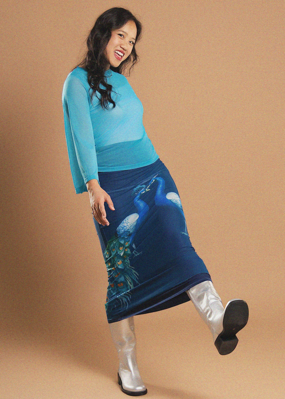 A woman modeling a two-piece outfit consisting of a sheer light blue long-sleeve top and a dark blue printed skirt with a peacock and jellyfish design, paired with silver boots, against a solid tan background.