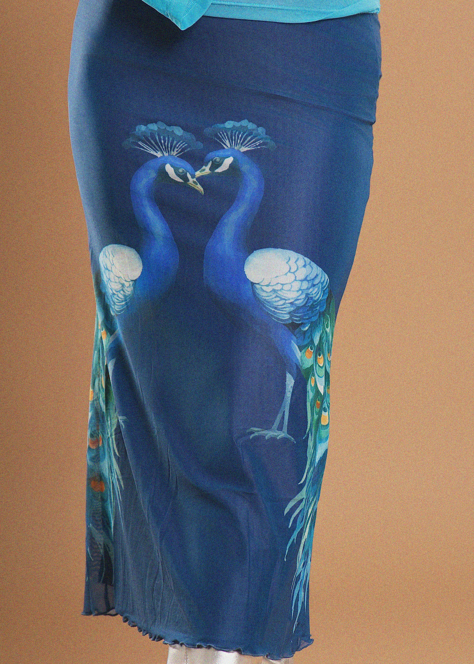 A blue skirt with a printed design of two peacocks facing each other, shown on a mannequin against a solid tan background.