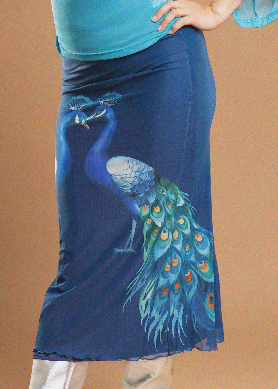 A blue midi skirt featuring a large printed peacock design with a vibrant green, blue, and orange tail, worn by a model against a solid tan background.
