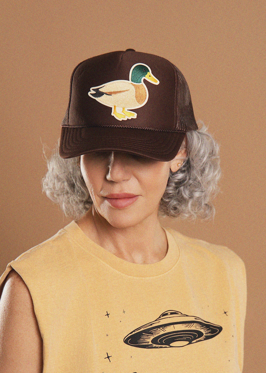 A woman with short gray curly hair wears a dark brown trucker hat featuring an embroidered mallard duck on the front. She is wearing a sleeveless beige top with a black UFO graphic, set against a solid warm brown background.