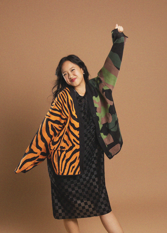A woman posing in a black dress with tiger-stripe patterned sleeves and a green camouflage patterned sleeve, set against a solid brown studio background.