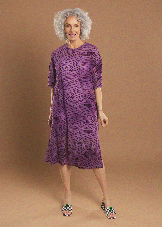 A woman with curly gray hair models a purple and black zebra-print short-sleeve dress against a solid tan studio background.