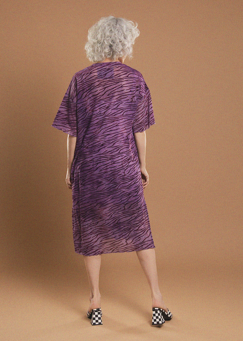 A woman with curly gray hair is shown from behind wearing a purple and black zebra-print short-sleeve dress, paired with black and white checkered heels, against a solid tan studio background.