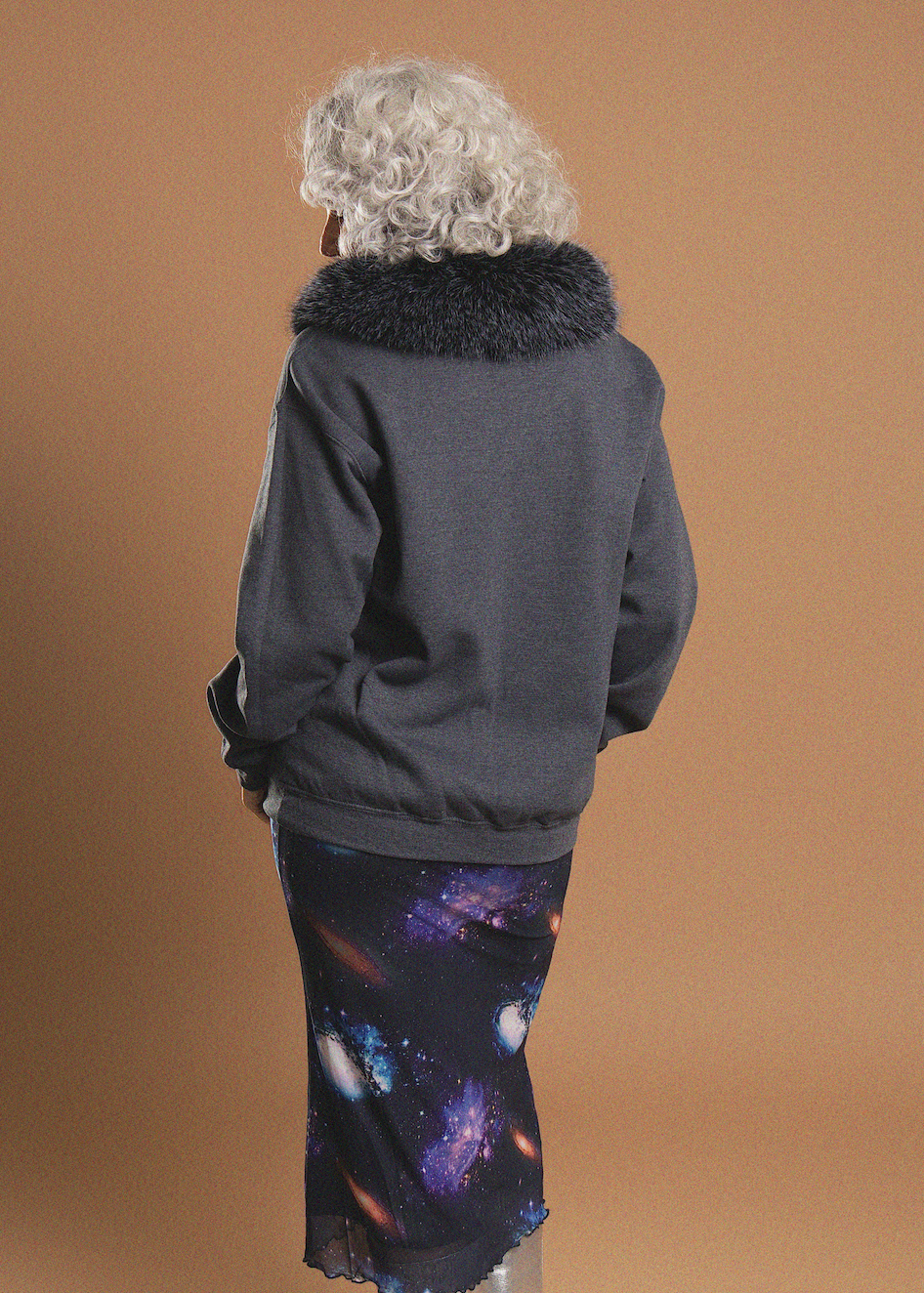 A person shown from the back wearing a dark gray jacket with a black faux-fur collar and a dark skirt with a colorful galaxy print, set against a solid tan studio background.