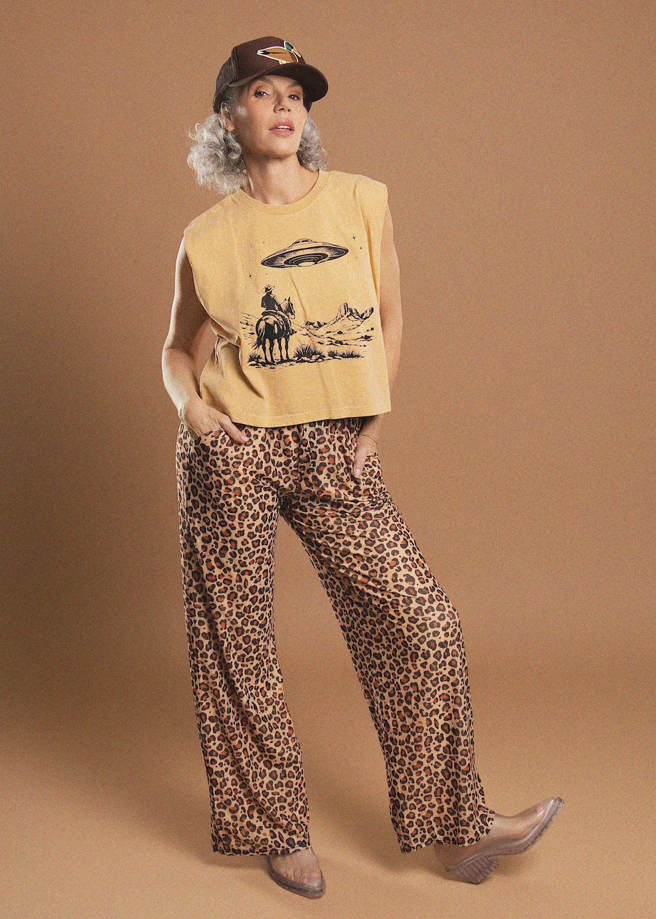 A model posing in a retro-style studio setting, wearing a tan sleeveless graphic t-shirt, leopard-print wide-leg pants, a brown baseball cap, and clear high-heeled boots against a solid tan backdrop.