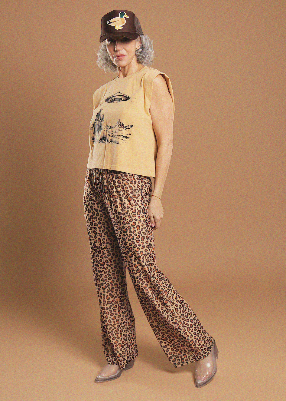 A woman modeling a sleeveless yellow top with a duck graphic, leopard-print pants, a brown duck cap, and clear pointed shoes against a solid tan studio background.