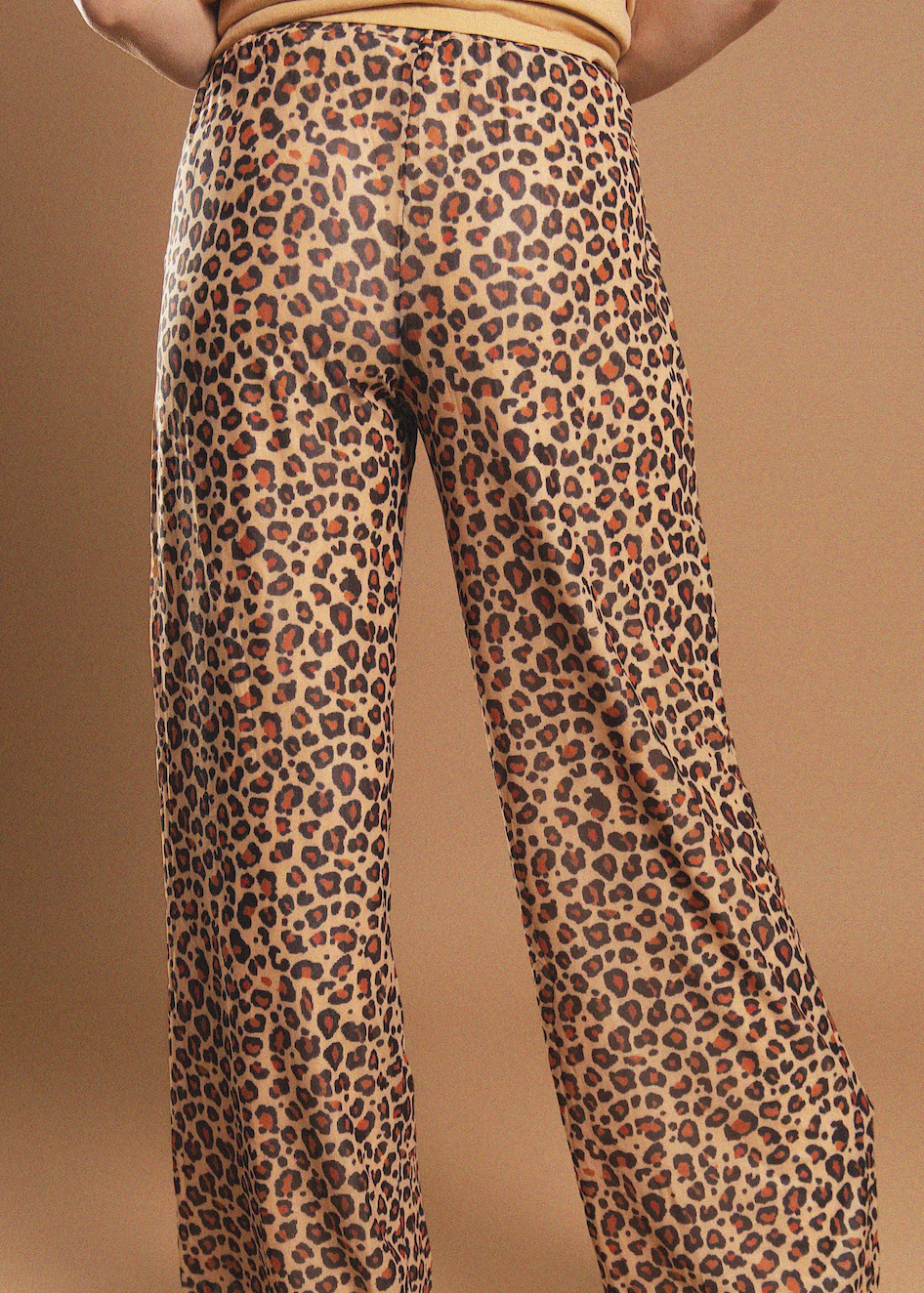Close-up back view of a person wearing leopard-print wide-leg pants against a warm tan studio background.
