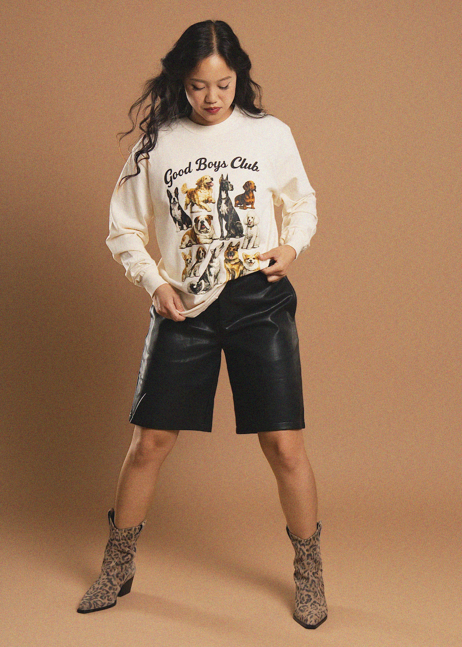 A woman modeling a cream sweatshirt with a 'Good Boys Club' dog graphic, paired with black faux-leather shorts and leopard-print ankle boots, against a solid tan studio background.