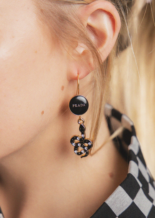A close-up of a model wearing a black circular Prada drop earring with a gold-tone hook and a dangling black and gold-tone floral charm embellished with small clear rhinestones.