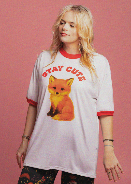 A woman wearing a white ringer t-shirt with red trim and a graphic of a fox with the text 'STAY CUTE' on the front, set against a solid pink background.