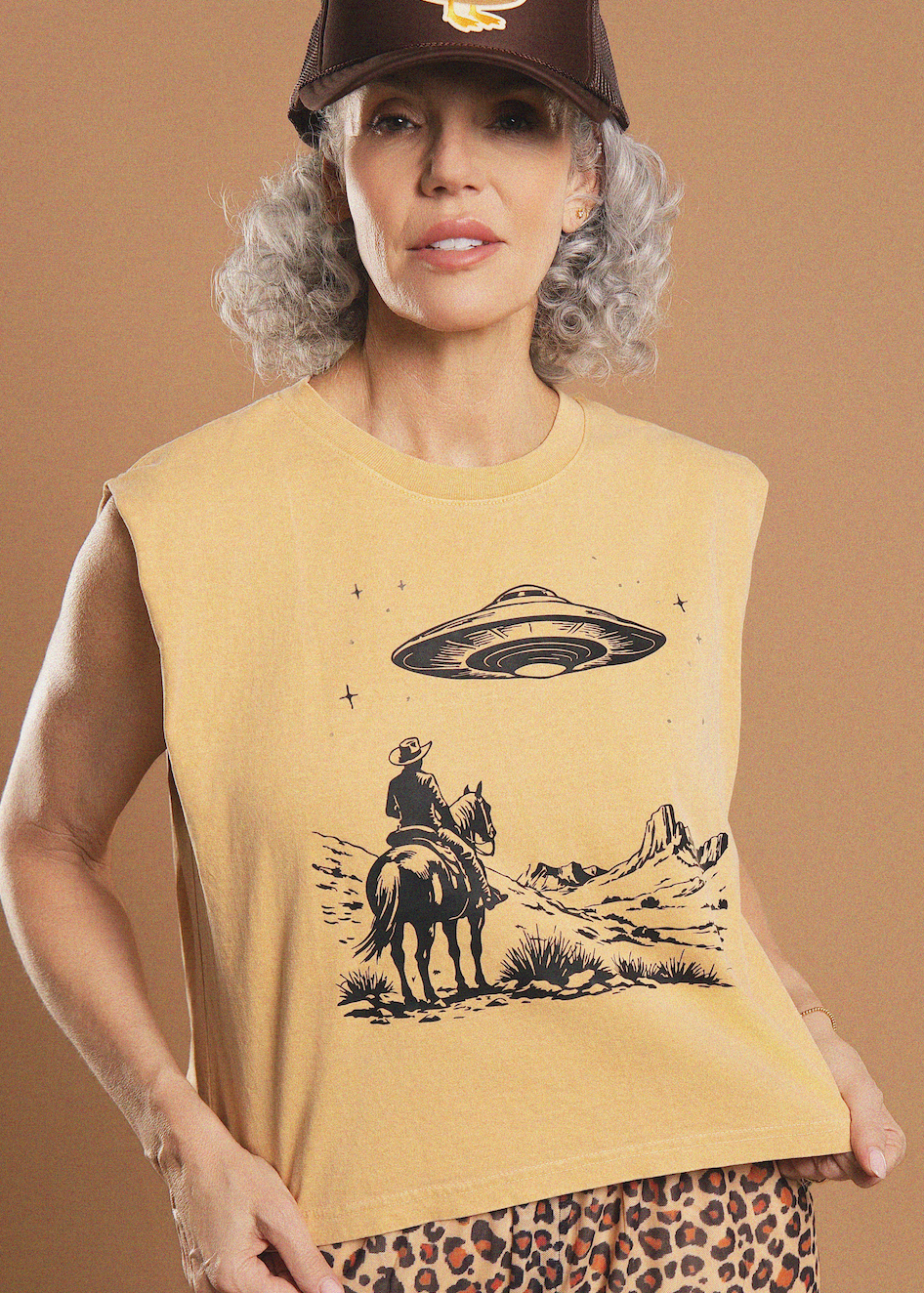 A woman wearing a tan sleeveless graphic t-shirt with a black print of a cowboy on a horse under a flying saucer, paired with a brown cap and leopard-print bottoms, set against a solid tan studio background.
