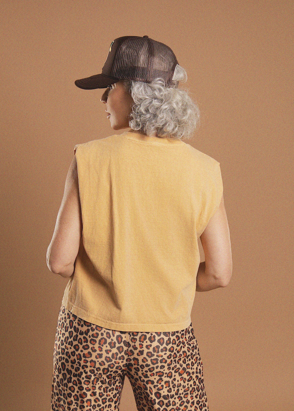 A person shown from the back wearing a mustard yellow sleeveless crop top, leopard-print pants, a dark trucker hat, and a curly gray wig, against a solid tan studio background.