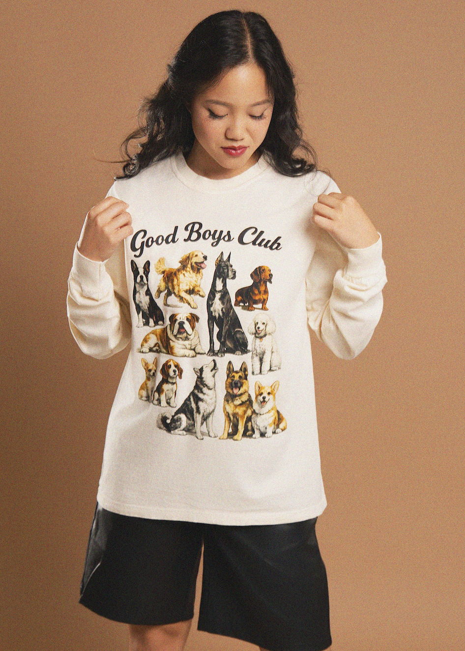 A young woman wearing a cream-colored long-sleeve t-shirt with a 'Good Boys Club' graphic featuring various dog illustrations, paired with black shorts, set against a solid tan studio background.