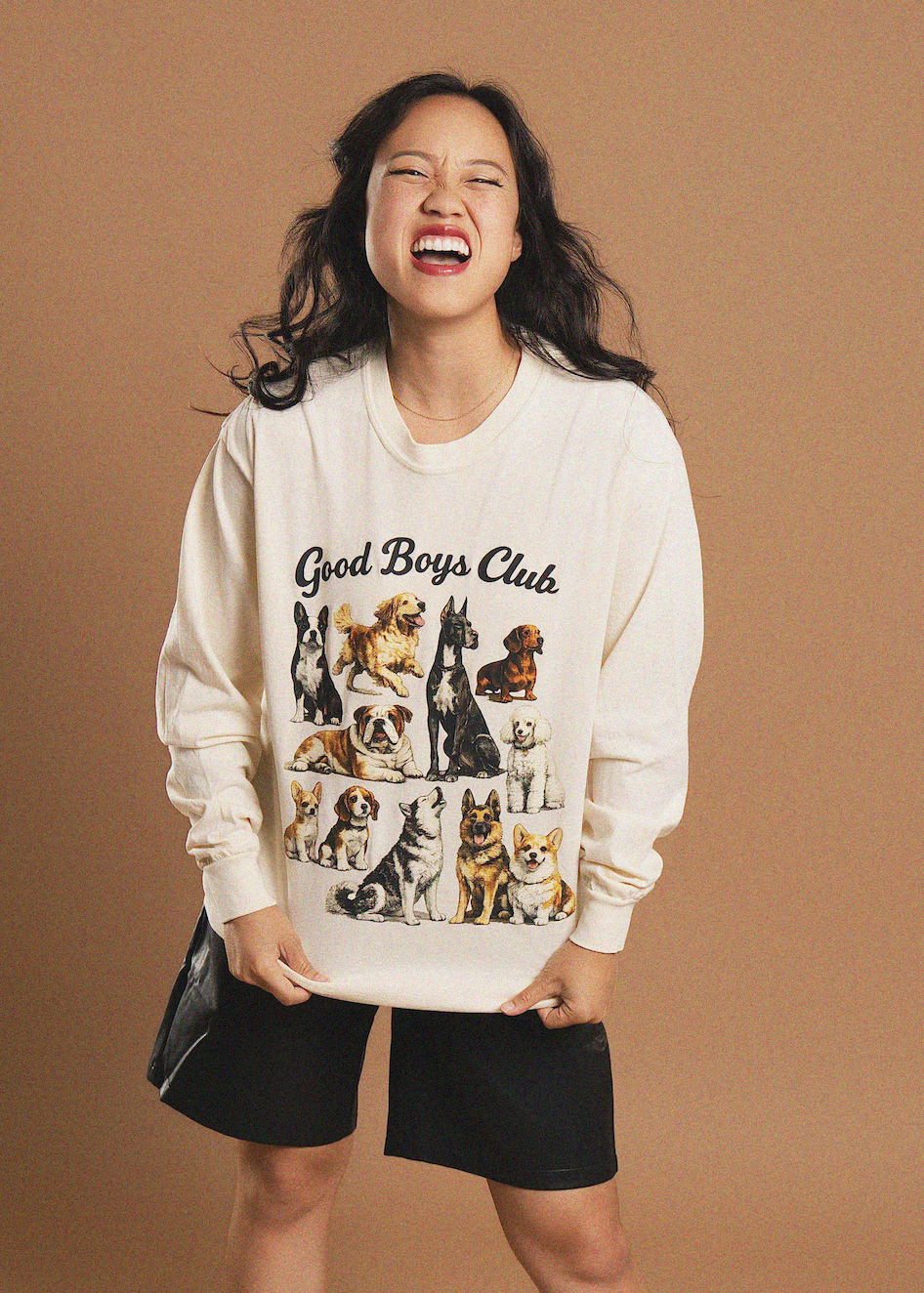 A woman laughing while wearing an off-white crewneck sweatshirt with a colorful graphic of various dog breeds and the text 'Good Boys Club', paired with black shorts, against a solid tan background.