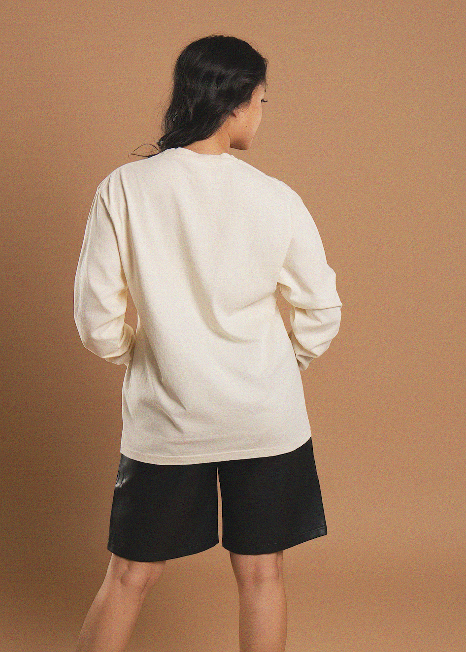 A model shown from the back wearing an oversized cream-colored long-sleeve shirt and black shorts, standing against a solid warm brown studio background.