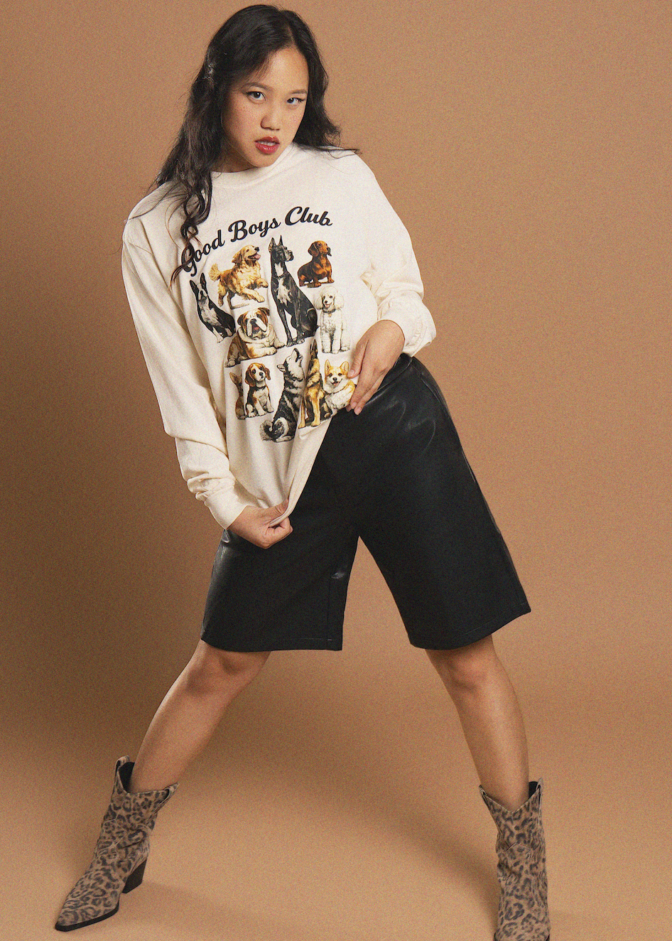 A model poses in a cream-colored graphic sweatshirt featuring a 'Good Boys Club' dog print, paired with black faux-leather shorts and leopard-print ankle boots, set against a solid tan studio backdrop.