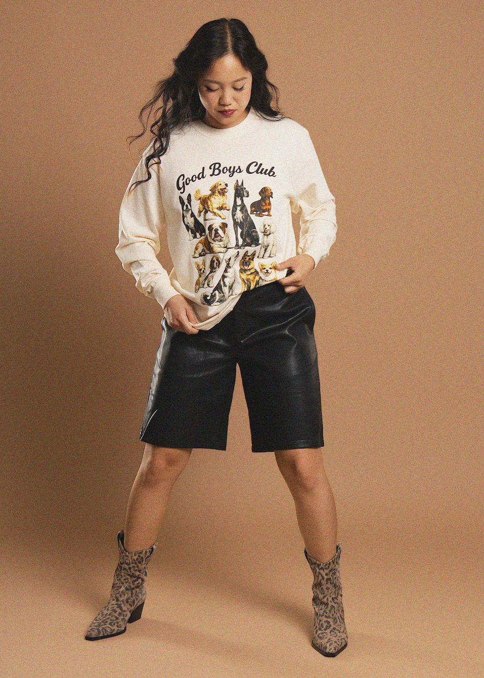 A woman modeling a cream sweatshirt with a 'Good Boys Club' dog graphic, paired with black faux-leather shorts and leopard-print ankle boots, against a solid tan studio background.