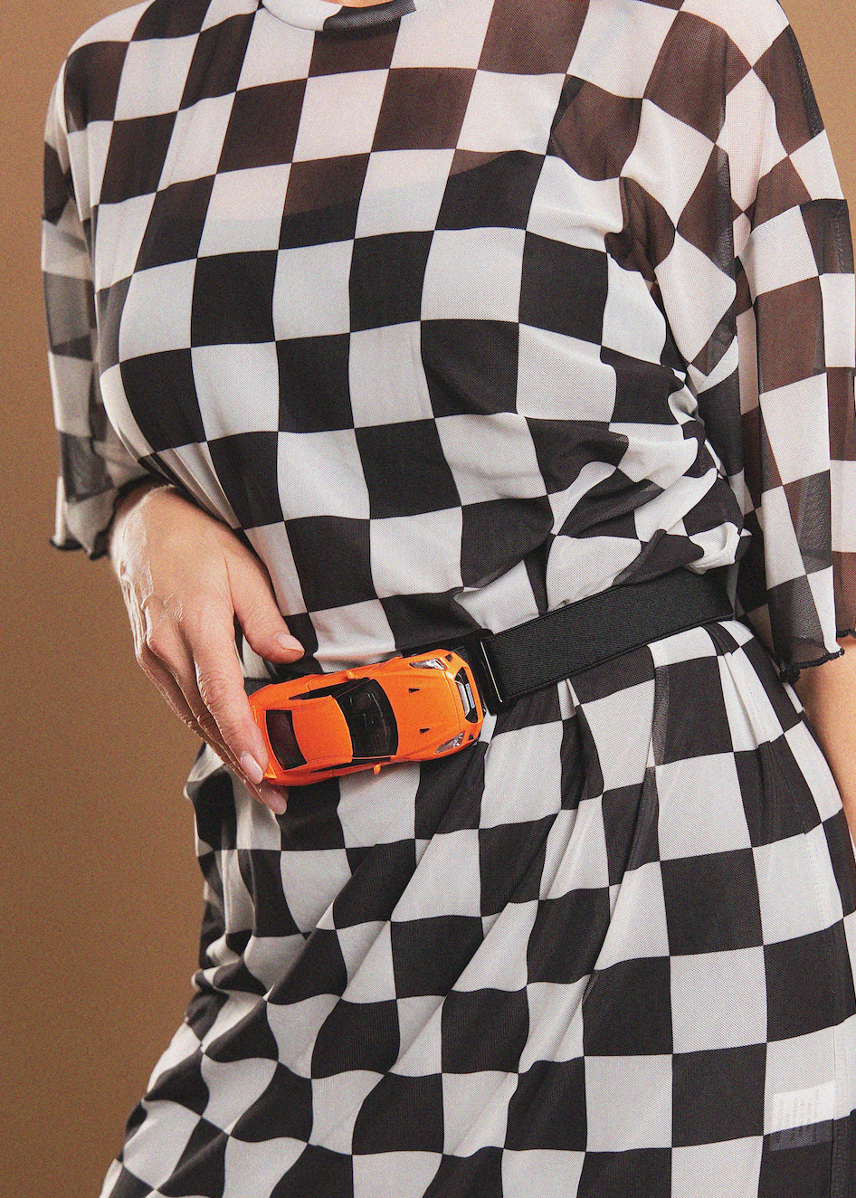 A close-up of a person wearing a black and white checkered dress and a black belt, holding an orange toy car in front of the belt.