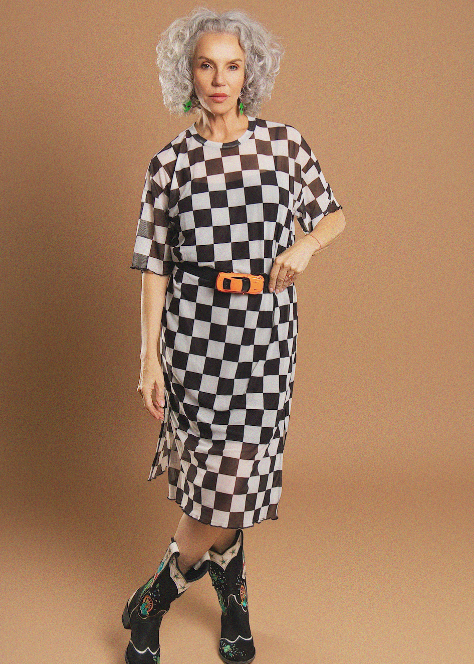 A woman modeling a short-sleeve midi dress with a black and white checkerboard pattern, styled with a black belt featuring a bright orange buckle. She is standing against a solid tan studio background.
