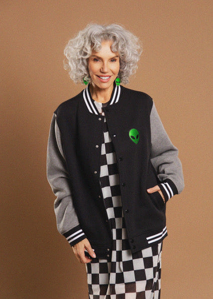 A woman with curly gray hair models a varsity jacket with a black and gray color-blocked design, featuring a green alien graphic on the chest. She is also wearing a black and white checkered dress against a solid brown studio background.
