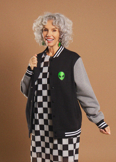 A woman with curly gray hair models a varsity jacket featuring a black body, gray sleeves, and a green alien head patch on the chest, worn over a black and white checkered dress against a solid brown studio background.