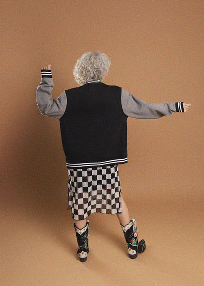 A model from behind wearing a varsity-style jacket with a black body and gray sleeves, paired with a black and white checkered skirt and western boots, set against a solid tan studio backdrop.