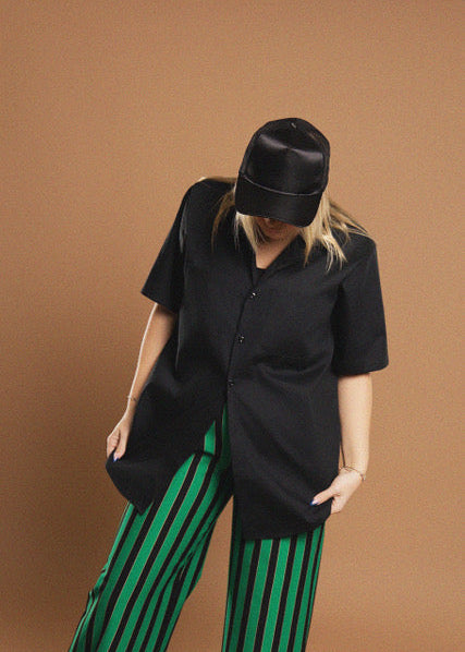 A model wearing a black oversized short-sleeve button-up shirt, a black cap, and green and black vertically striped pants, posed against a solid warm brown studio background.