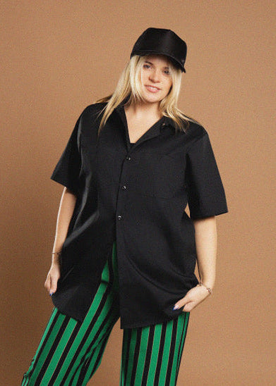 A woman modeling a black short-sleeve button-up shirt and black cap against a solid brown studio background, paired with green and black striped pants.