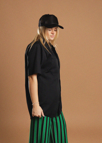 A woman wearing a black short-sleeve button-up shirt, a black baseball cap, and a green and black vertically striped skirt, standing against a solid brown studio background.