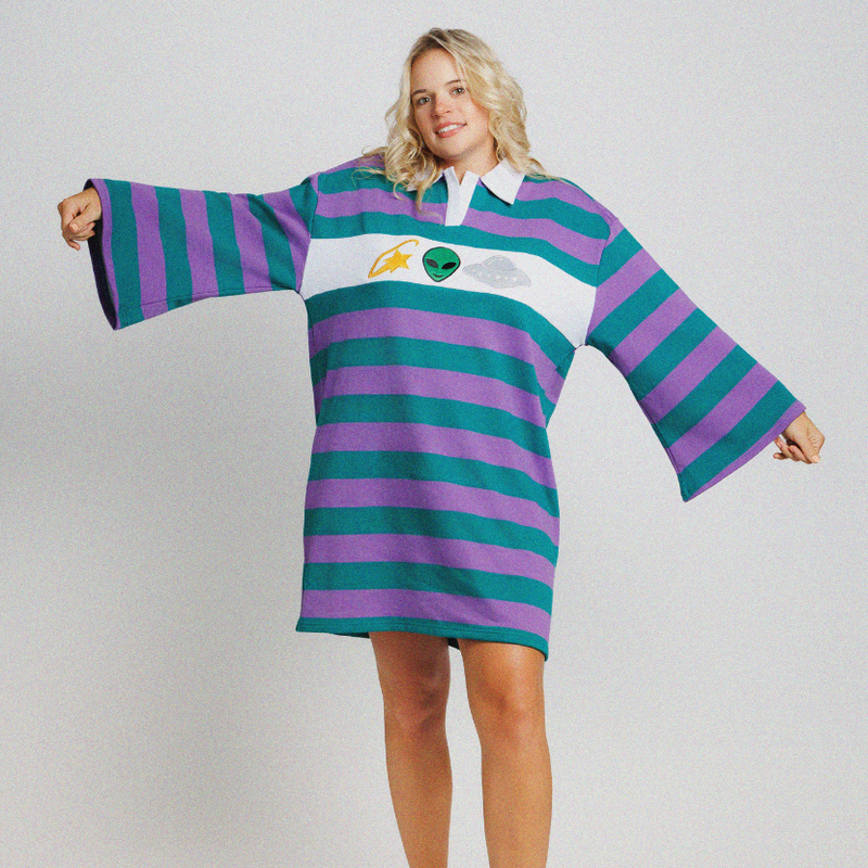 A woman modeling a long-sleeve rugby-style mini dress with bold horizontal purple and teal stripes, a white collar, and a green chest emblem, photographed against a light gray studio background.