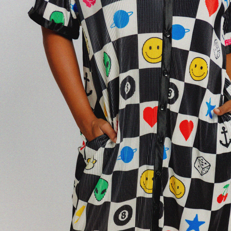 A person wearing a short-sleeve maxi dress with a black and white checkerboard pattern decorated with colorful smiley faces, hearts, stars, and other playful symbols, shown against a plain light gray background.