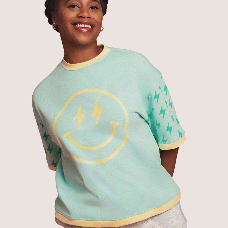 A smiling woman wearing a mint green ringer t-shirt with a yellow smiley face graphic and yellow trim, set against a solid pale pink background.